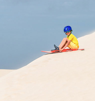 A kid wearing a helmet enjoys sandboarding at a School Camp of South Gates beach, Geraldton Fairbridge Camp